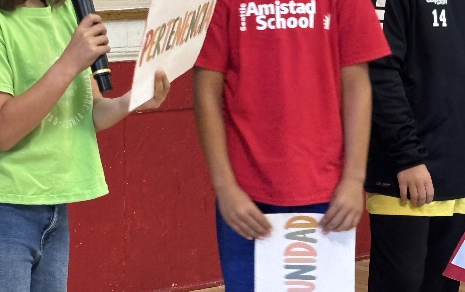 Seattle Amistad 4th and 5th graders stand in a line in the gym holding signs with Spanish words including Escucha, Ayuda, Curiosidad, and Paciencia.