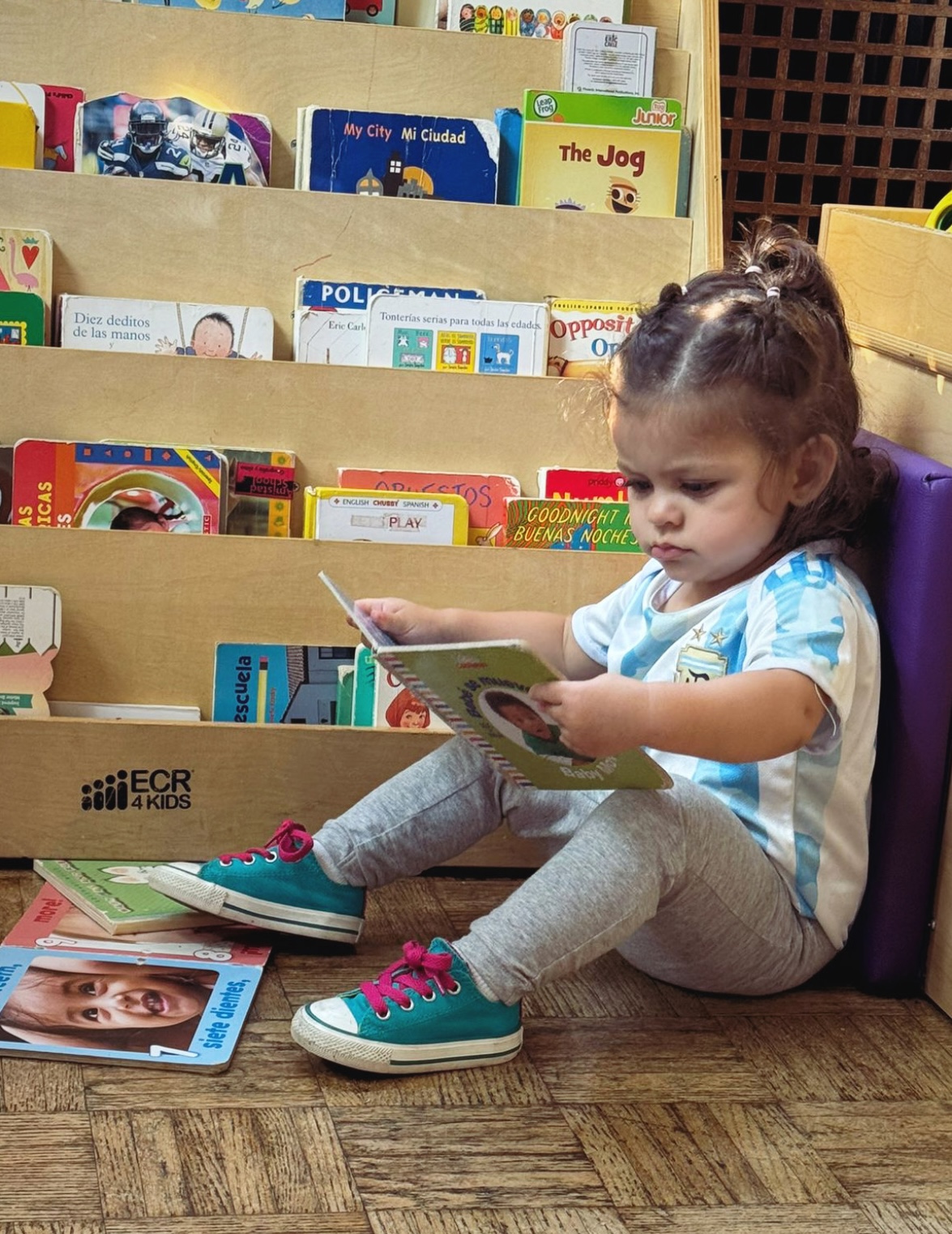 A toddler sitting by the bilingual book shelf turning the pages of a board book