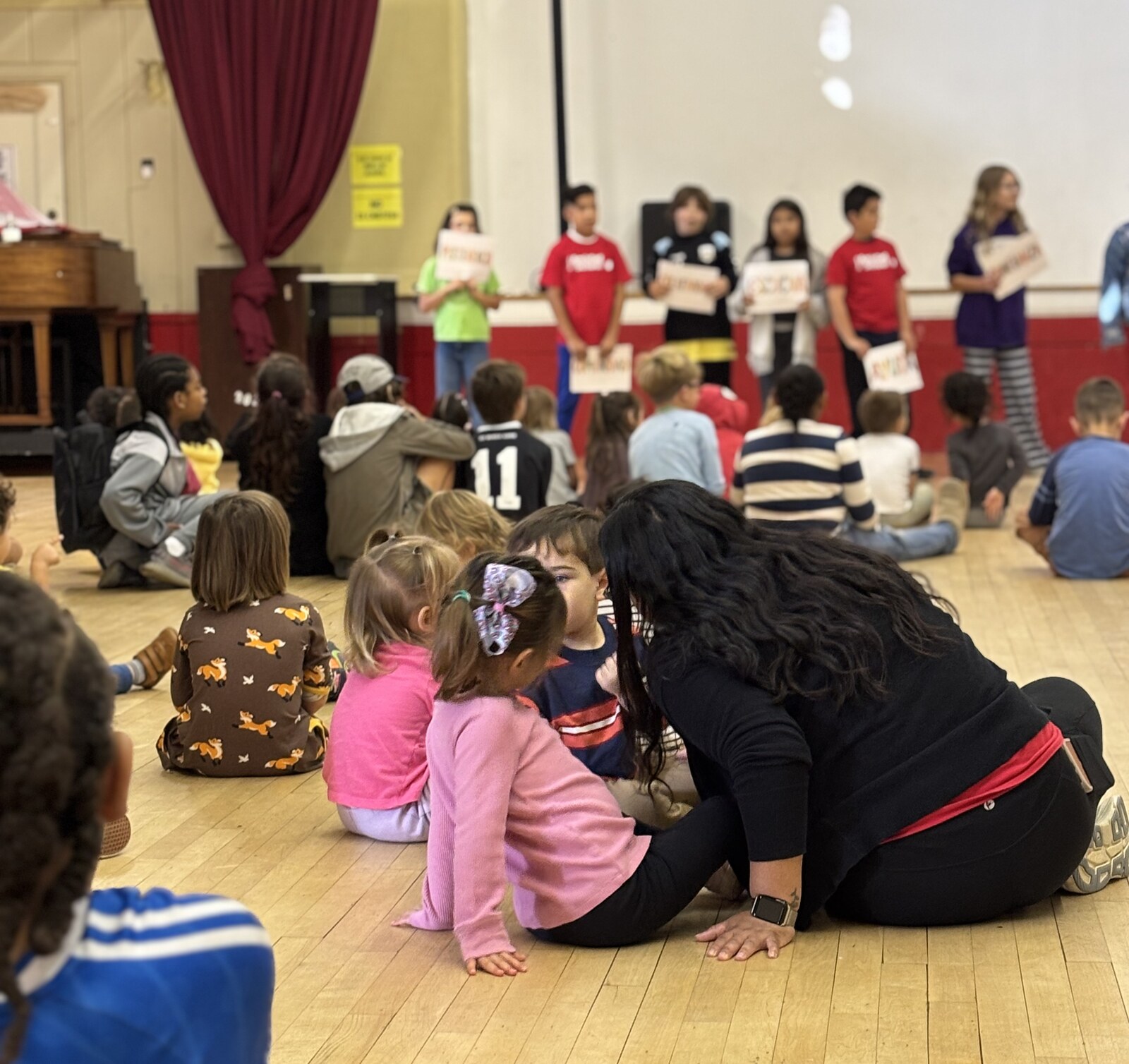 Younger students sit on the gym floor watching older students hold word signs during Hora de Amistad.